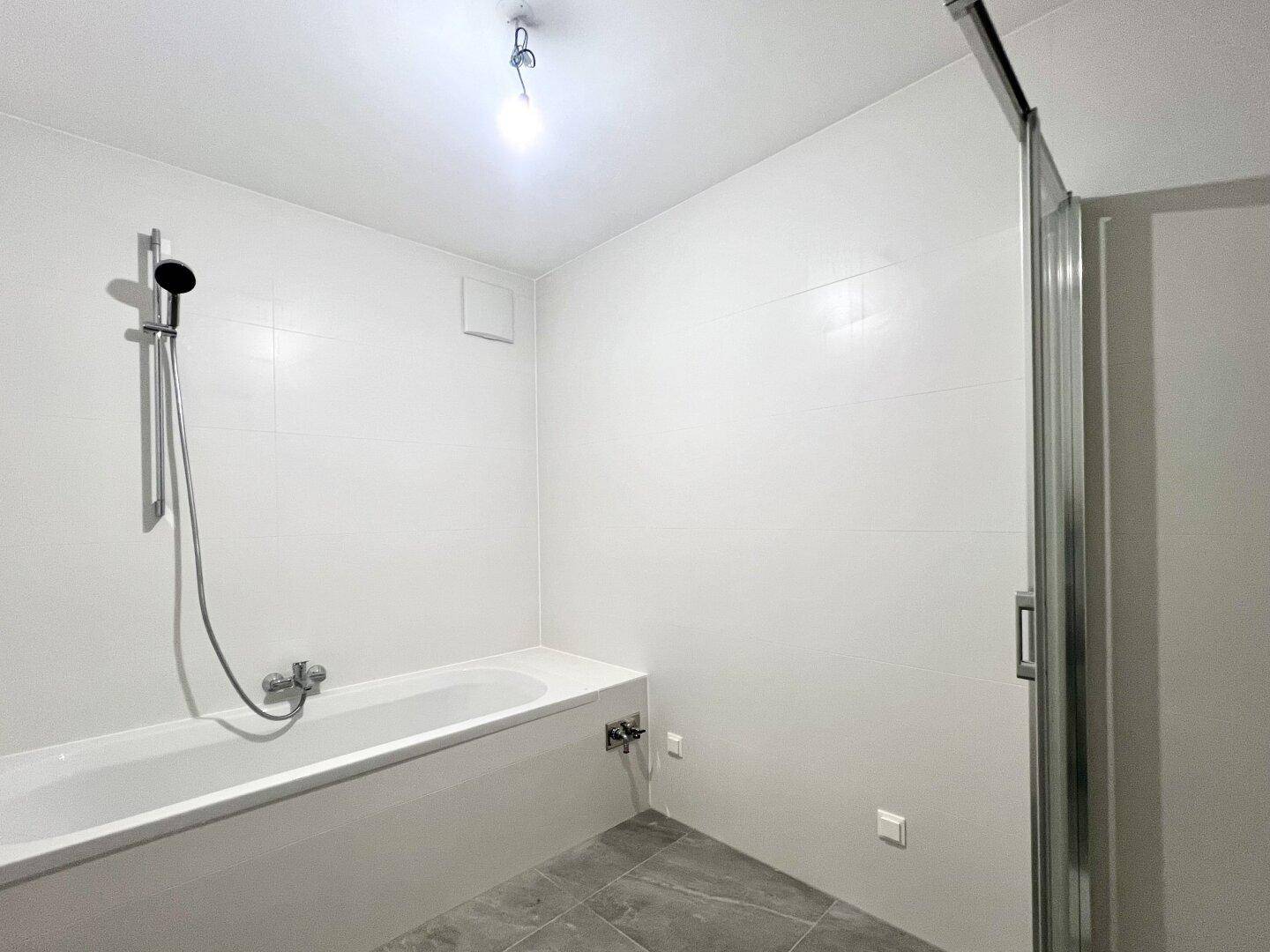Modernes Badezimmer mit weißen Fliesen, grauen Bodenfliesen, einer Badewanne und einer Duschabtrennung.
