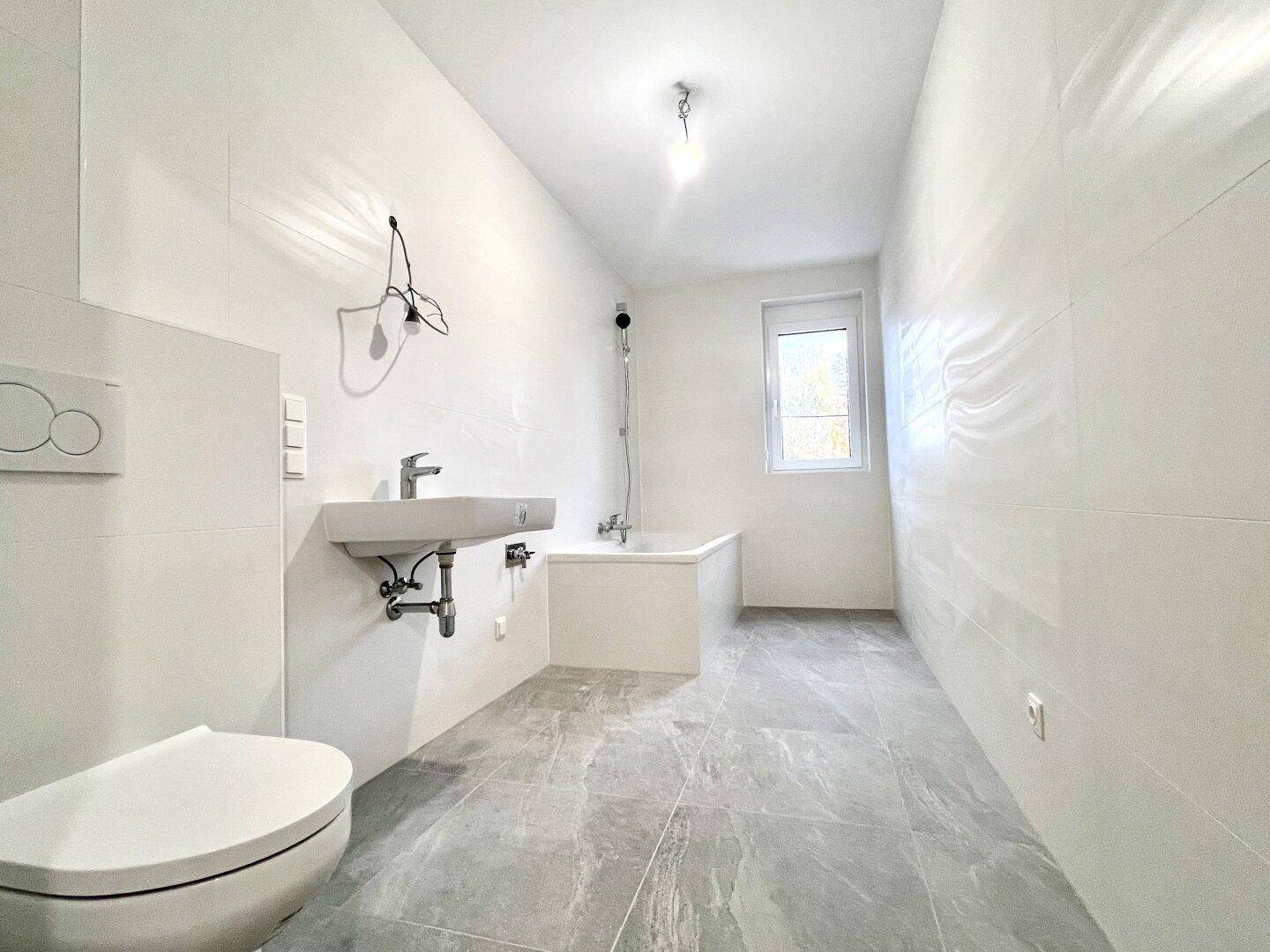 Modernes Badezimmer mit weißen Fliesen, grauen Bodenfliesen, Badewanne, Waschbecken und WC.