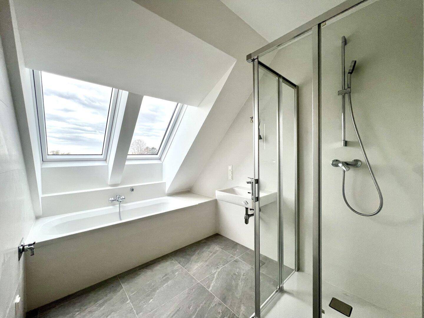 Helles Badezimmer mit Badewanne, Duschkabine und modernen grauen Bodenfliesen im Dachgeschoss.