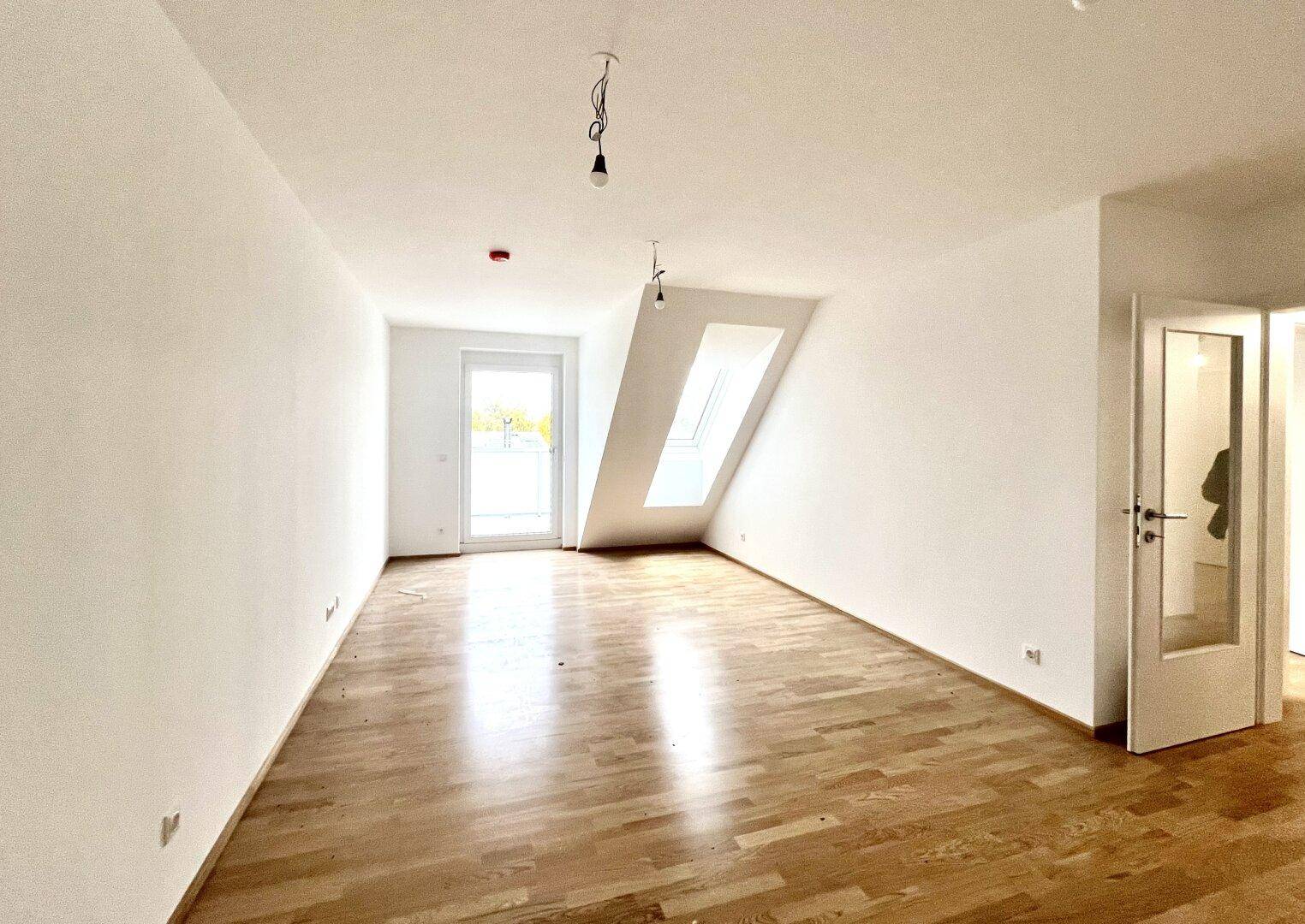 Helles Zimmer mit Holzboden, Dachschräge und Zugang zu einem Balkon im Dachgeschoss.