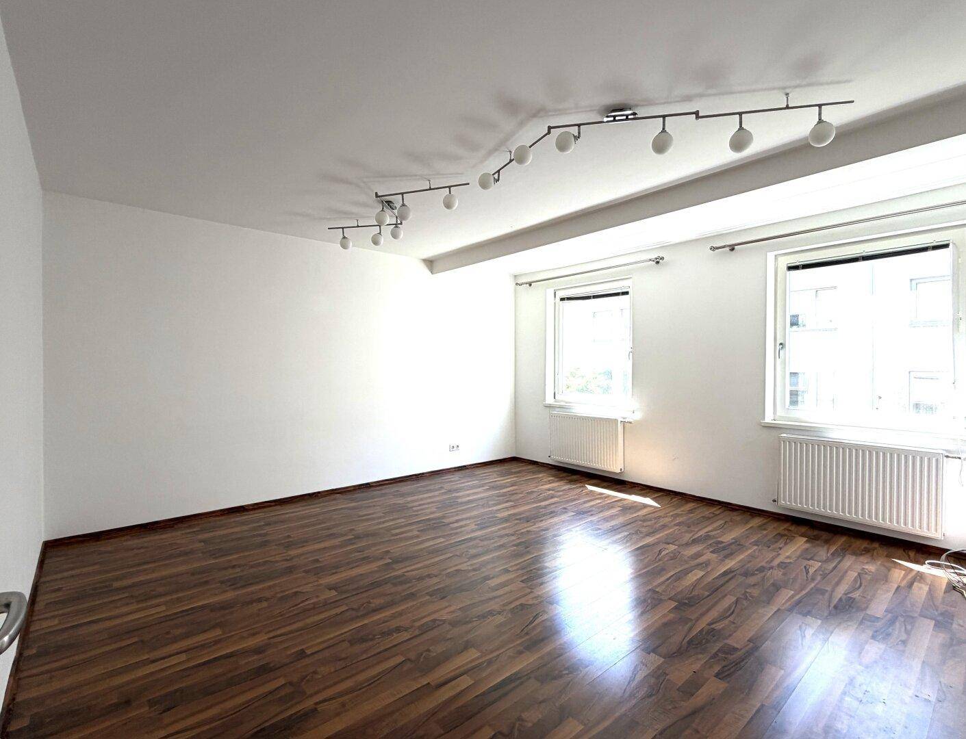 Helles, leeres Zimmer mit Holzboden, weißen Wänden und zwei Fenstern mit Heizkörpern.