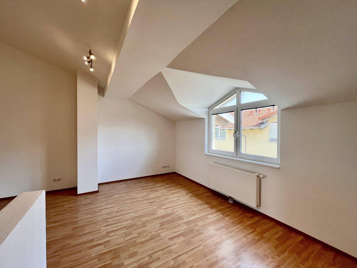 Heller Dachgeschossraum mit Holzboden, Heizkörper und Fenster mit Blick auf die Nachbarschaft.