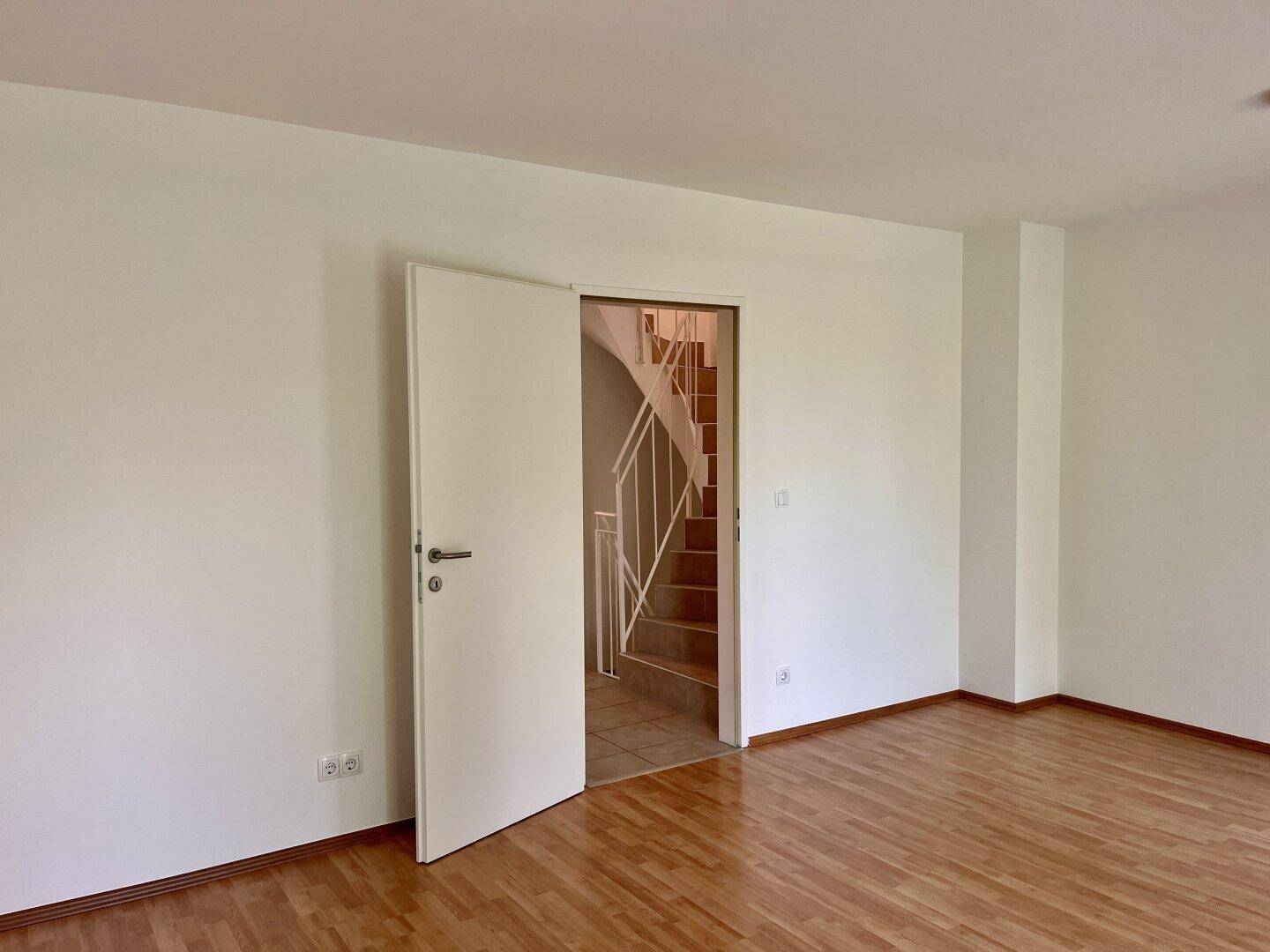 Leeres Zimmer mit Holzboden und offener Tür, die den Blick auf die Treppe freigibt.
