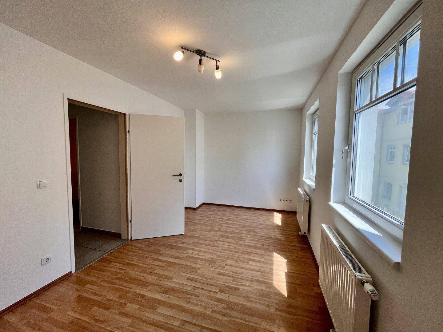 Helles Zimmer mit Holzboden, großen Fenstern und Heizkörpern, bereit zur Einrichtung.