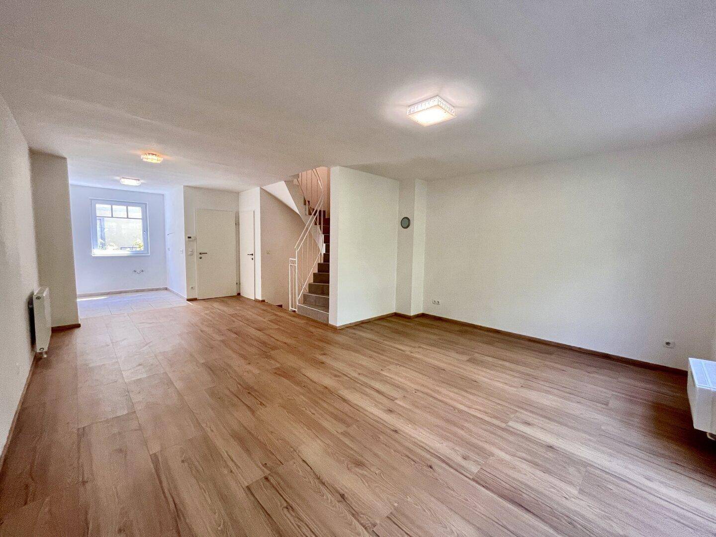 Geräumiges Wohnzimmer mit hellem Holzboden und Zugang zu einem weiteren Bereich sowie einer Treppe.
