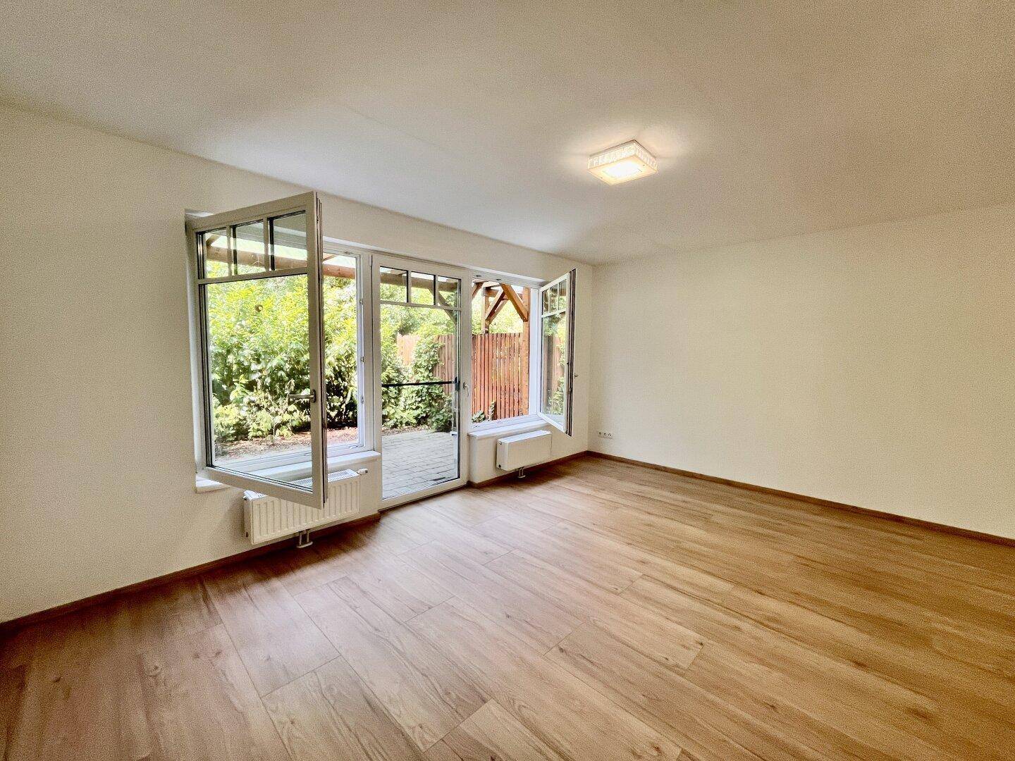 Heller Wohnraum mit Holzboden und geöffneten Fenstern, die den Blick in den Garten freigeben.