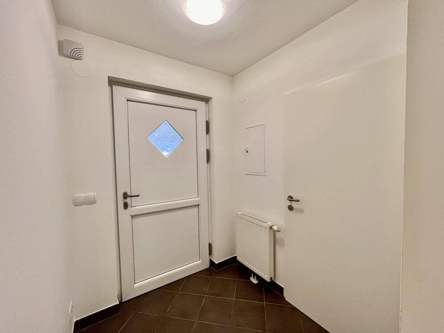 Eingangsbereich mit weißer Tür, dunklen Fliesen und einem Heizkörper an der Wand.