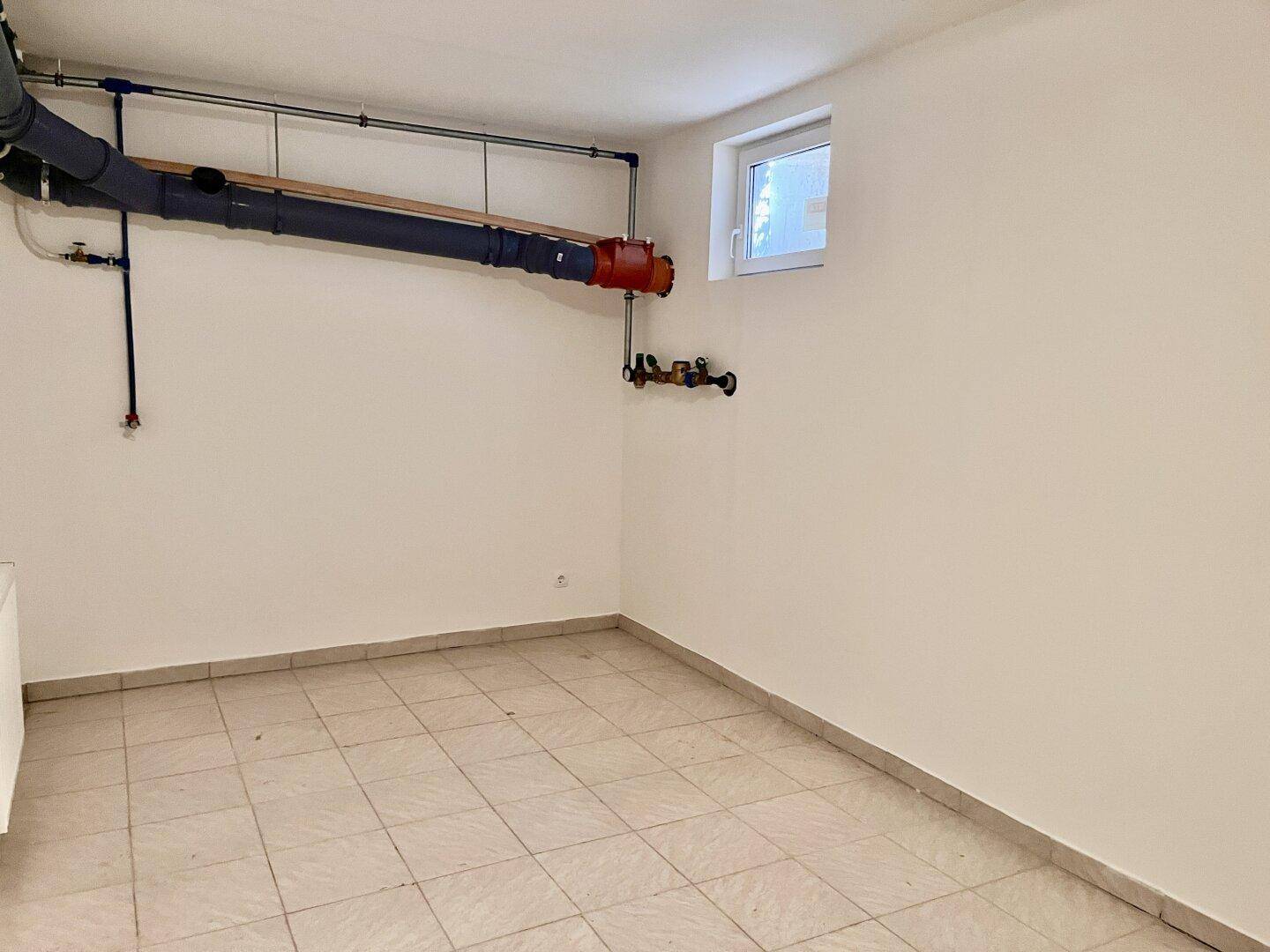 Hauswirtschaftsraum im Keller mit Fliesenboden, sichtbaren Leitungen und kleinem Fenster.