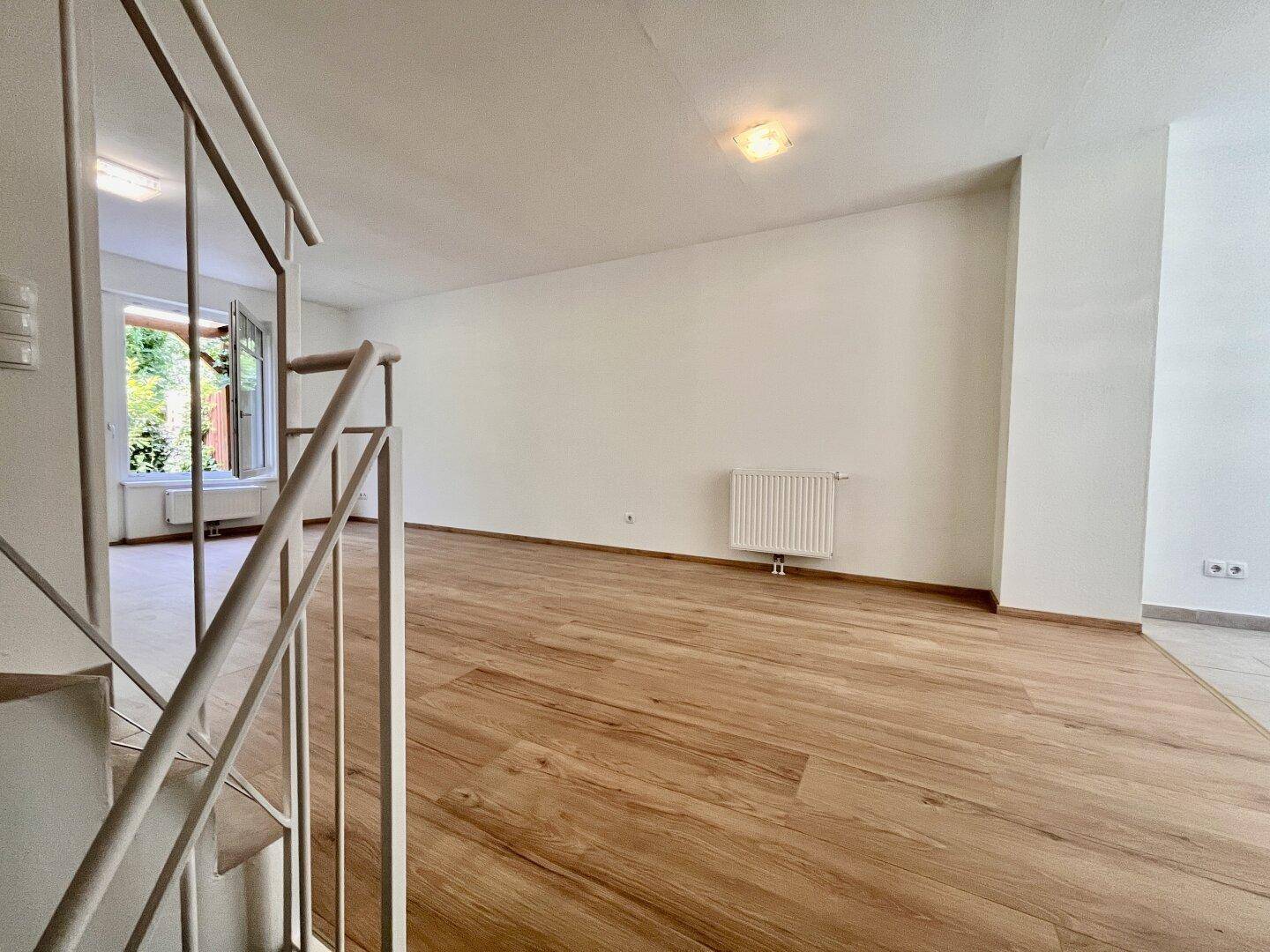 Blick vom Treppenaufgang in das helle Wohnzimmer mit Holzboden und großen Fenstern.