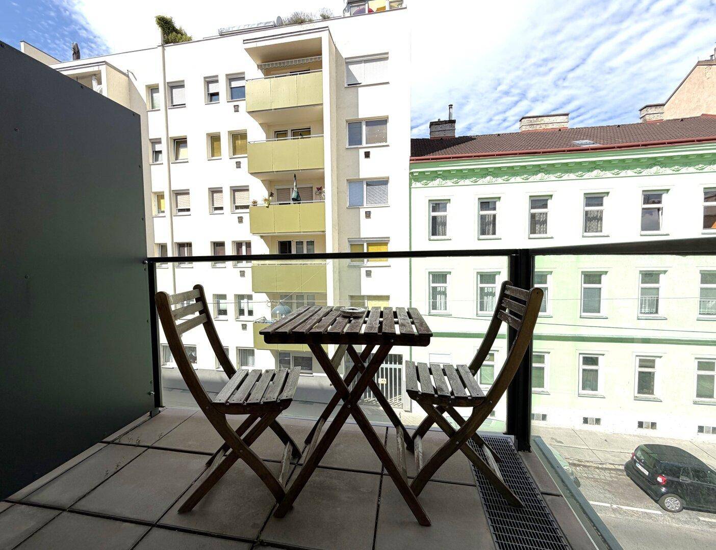 Kleiner Balkon mit Holztisch und Stühlen, bietet einen Blick auf die umliegenden Gebäude und die Straße.
