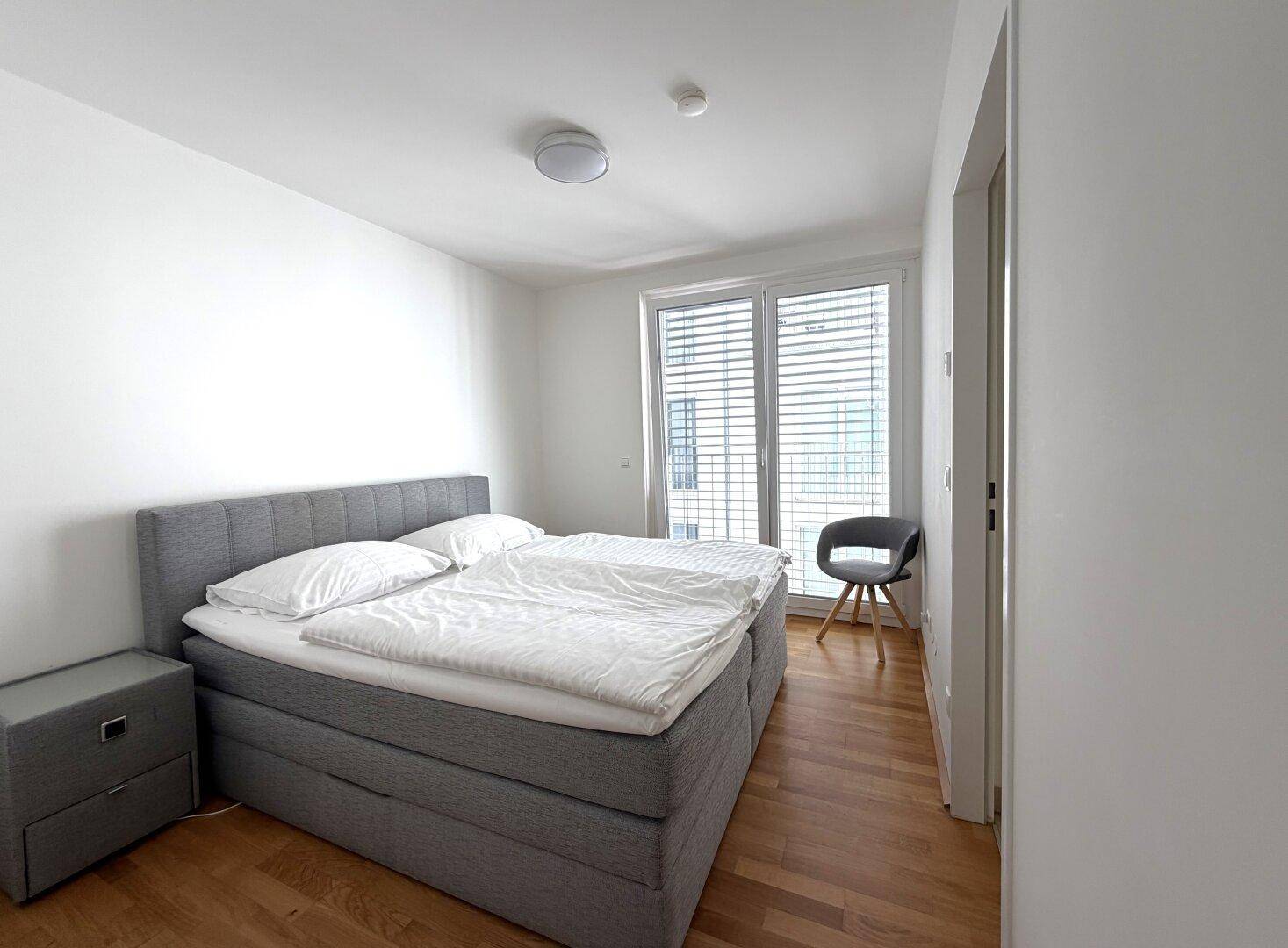 Helles Schlafzimmer mit Doppelbett, Nachttisch und großem Fenster mit Jalousien.