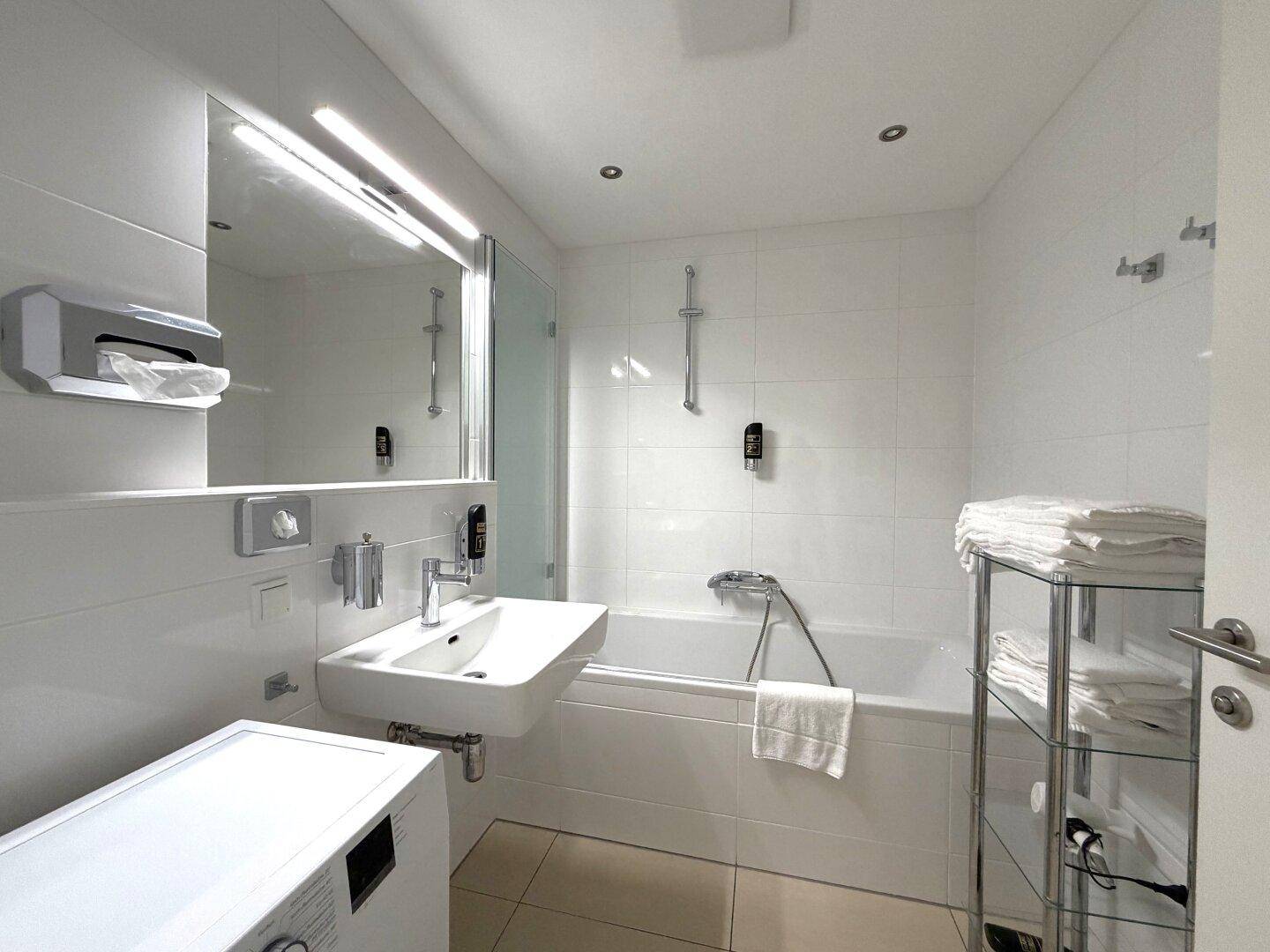 Modernes Badezimmer mit weißen Fliesen, Badewanne mit Duschfunktion und Waschmaschine.