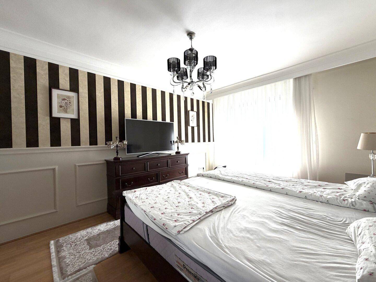 Geräumiges Schlafzimmer mit klassischem Holzbett, Kommode mit Fernseher und gestreifter Wand.
