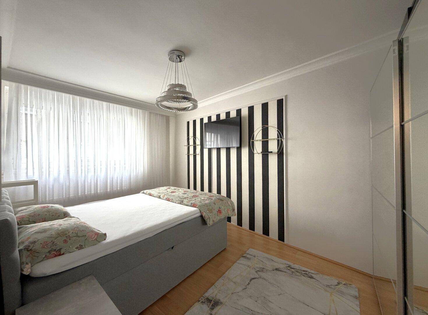 Helles Schlafzimmer mit Doppelbett, Holzfußboden und großen Fenstern mit transparenten Vorhängen.
