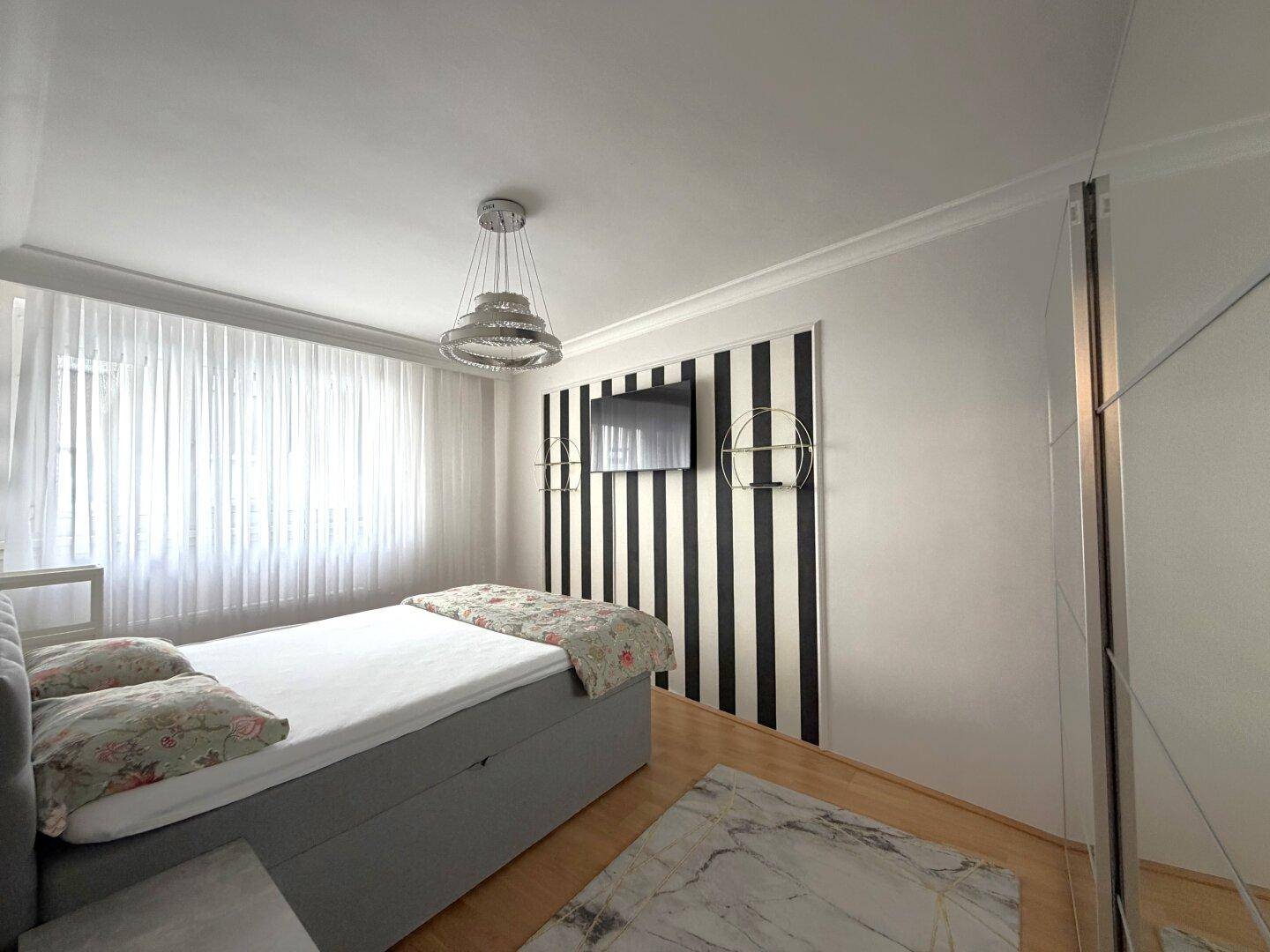 Schlafzimmer mit modernem Design, Wandfernseher und viel Tageslicht durch das große Fenster.
