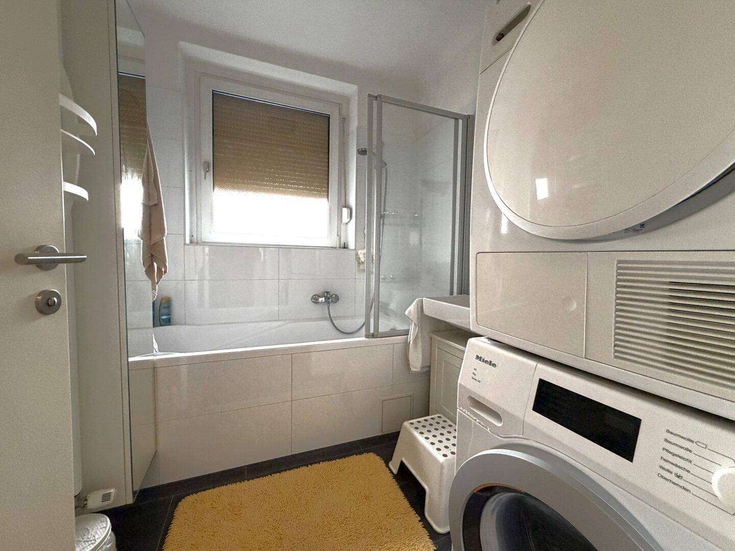 Modernes Badezimmer mit Badewanne, Duschwand, Fenster und integrierter Waschmaschine und Trockner.