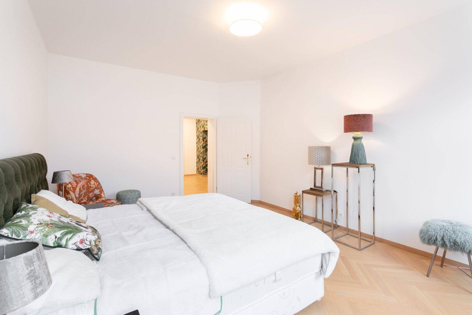 Helles Schlafzimmer mit Fischgrätparkett und einer Tür, die zu einem farbenfrohen Flur führt.