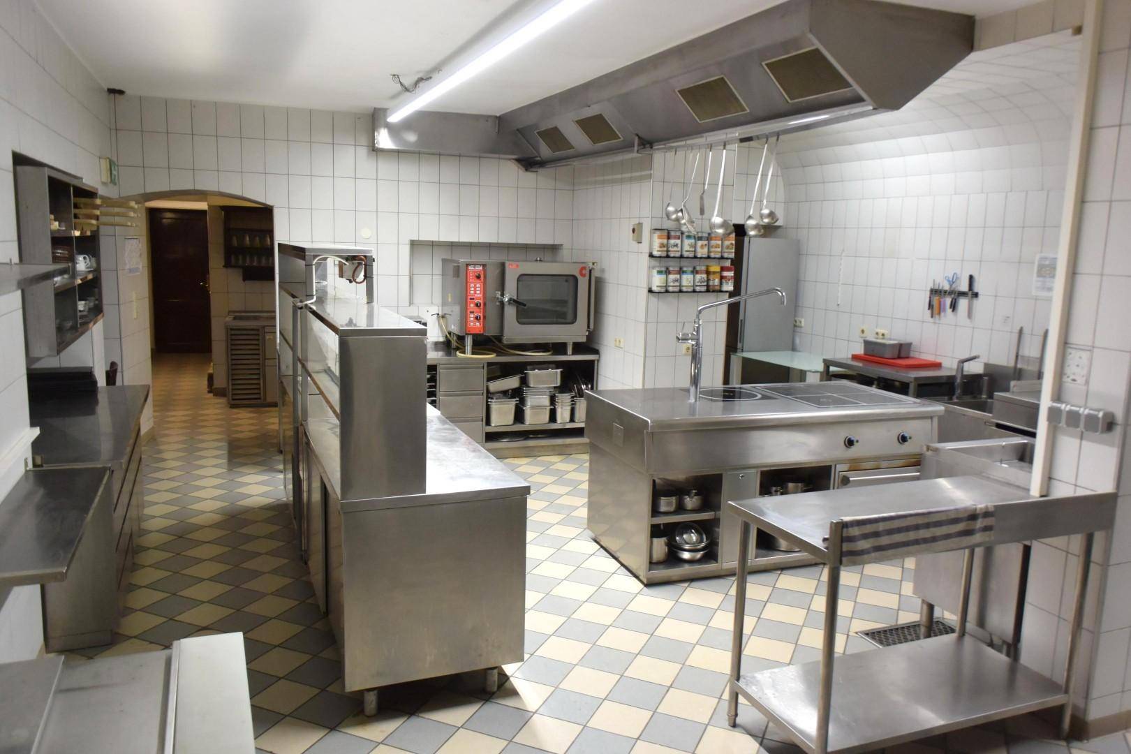 Große, professionelle Gastroküche mit Edelstahlgeräten, Fliesenwänden und einem karierten Fliesenboden.