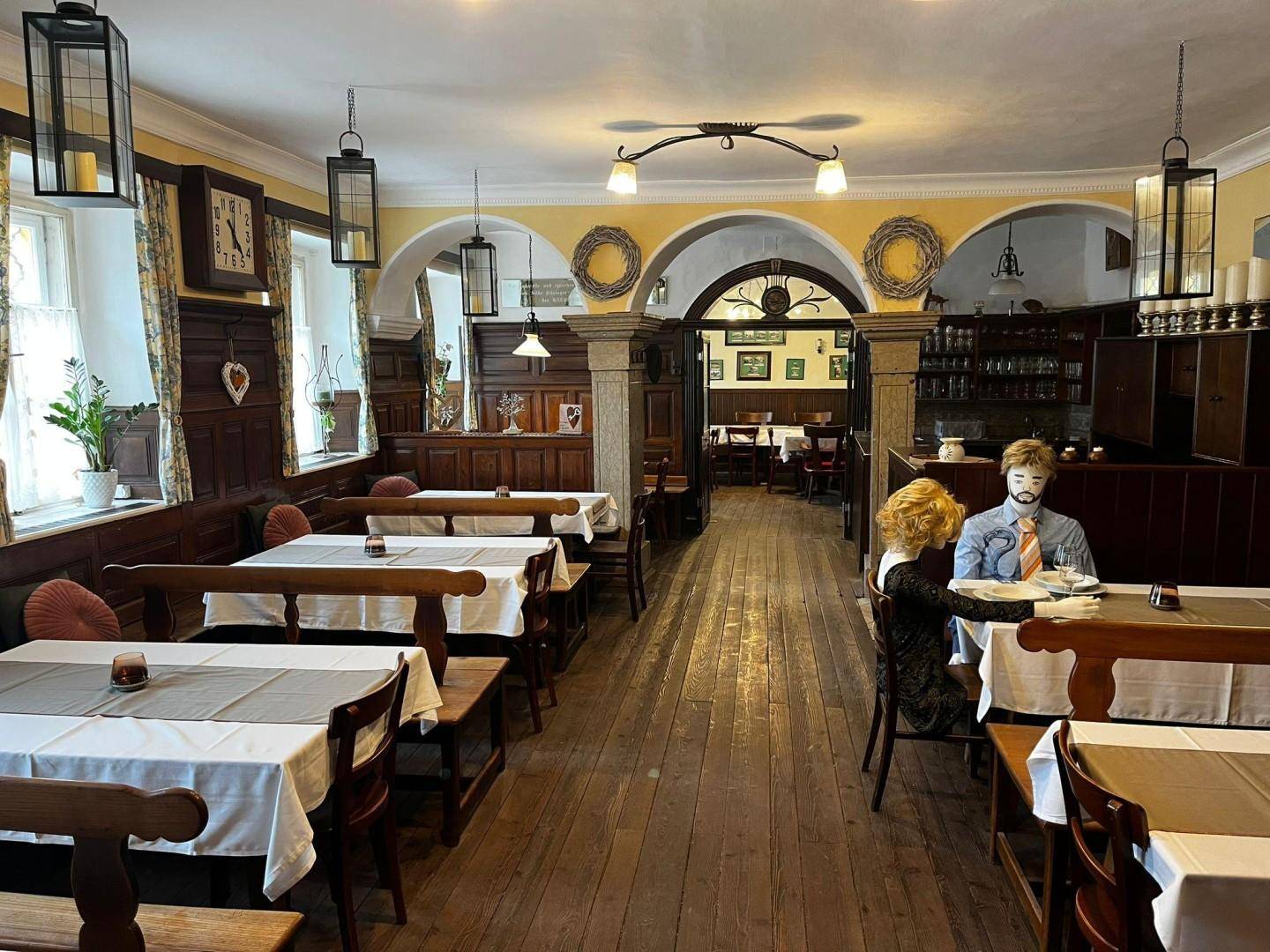 Geräumiger Gastraum mit Holzböden, Holzbänken, Tischen mit weißen Decken und dekorativen Wandkränzen.