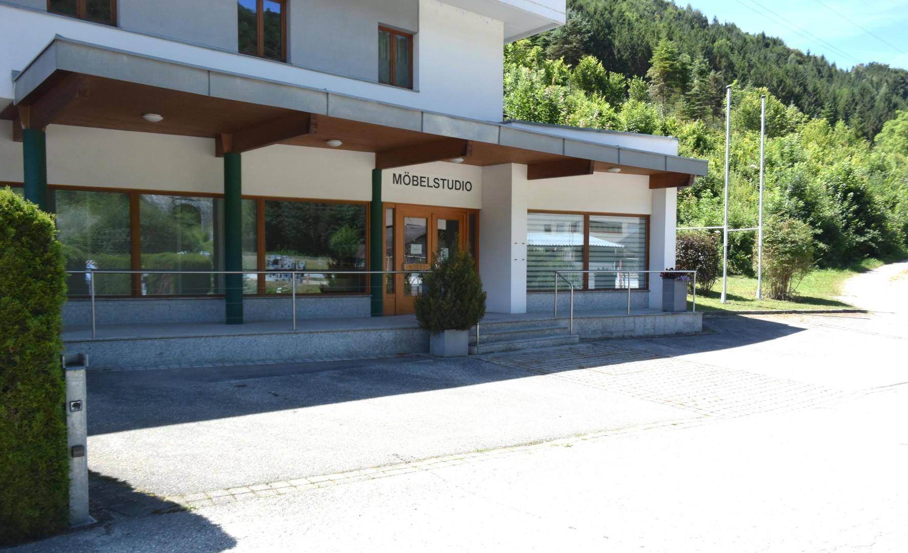 Modernes Geschäftsgebäude mit großen Glasfronten und einem Vordach, umgeben von grüner Landschaft und Bergen.
