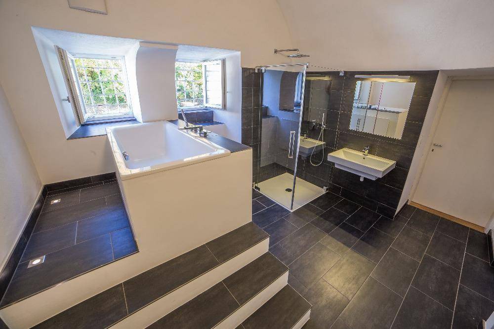 Geräumiges modernes Badezimmer mit erhöhter Badewanne, Glasdusche und dunklem Fliesenboden.