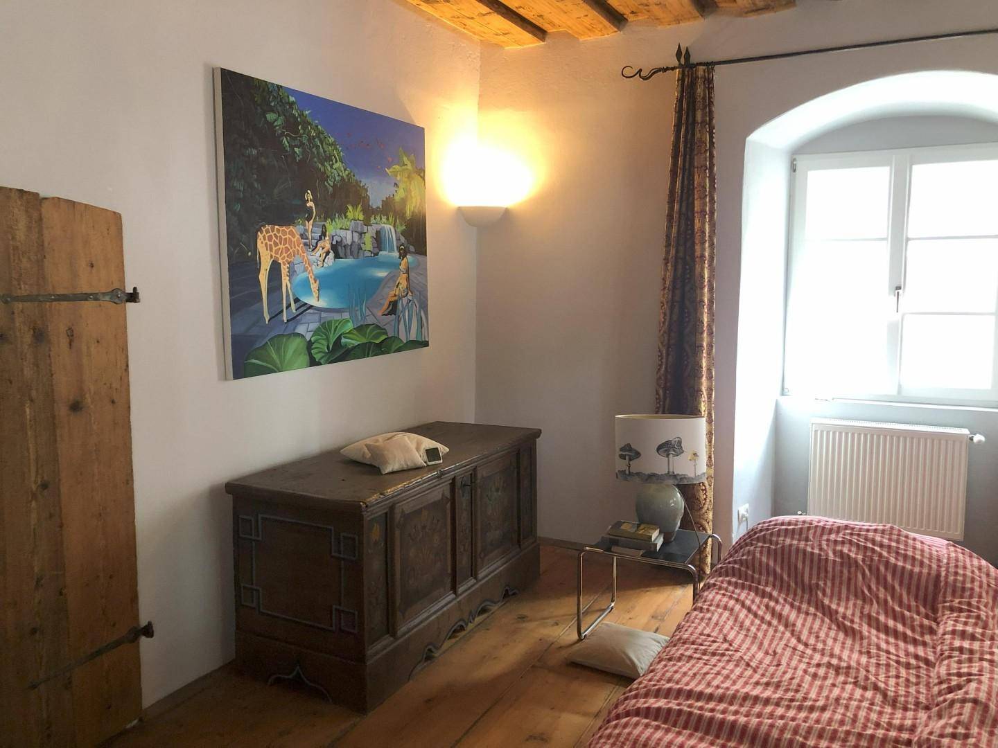 Schlafzimmer mit Holzbalkendecke, antiker Truhe und farbenfrohem Kunstwerk an der Wand.