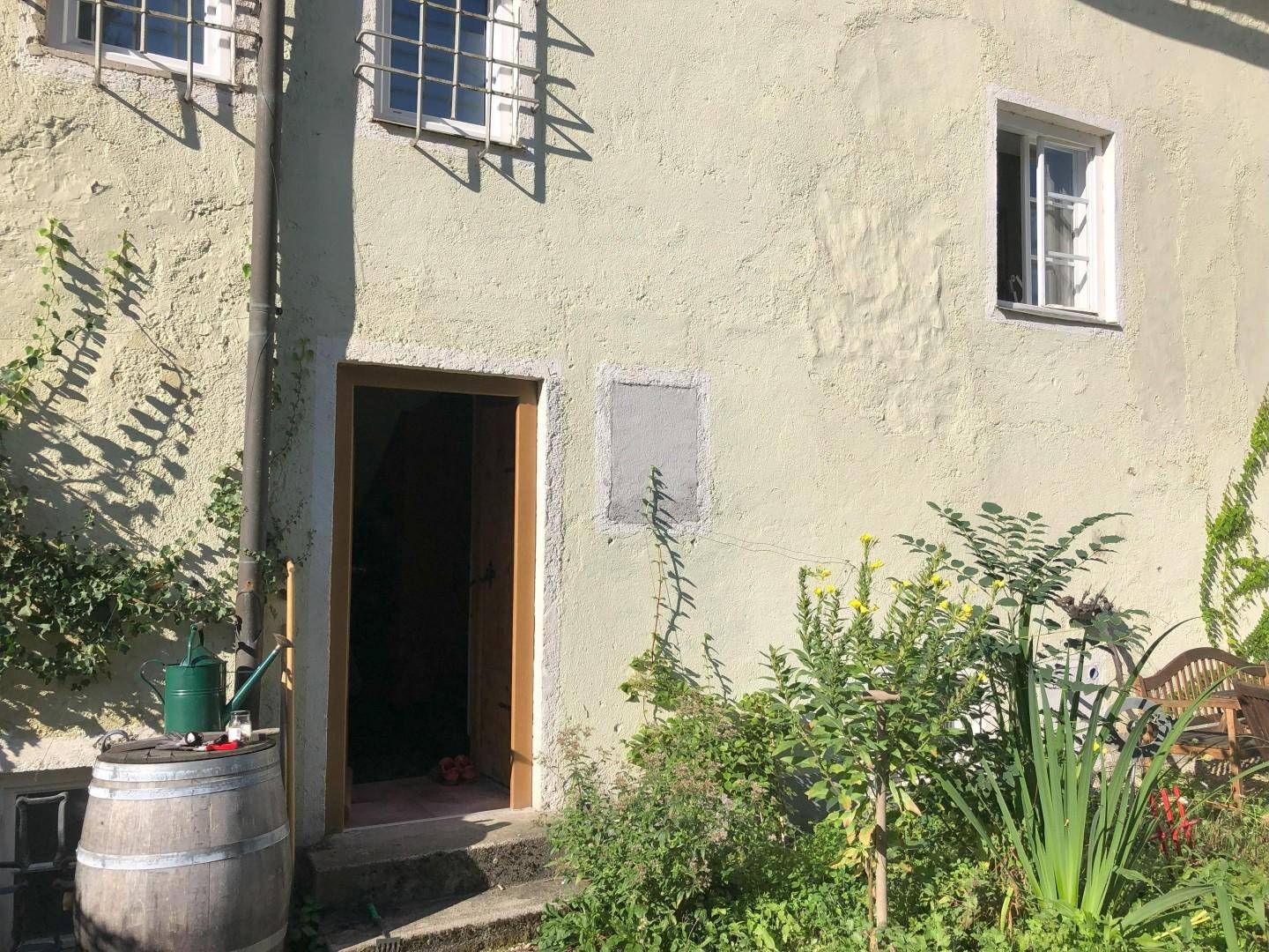 Seitlicher Eingang des Gebäudes, umgeben von einem grünen Garten mit Pflanzen und Gartenutensilien.