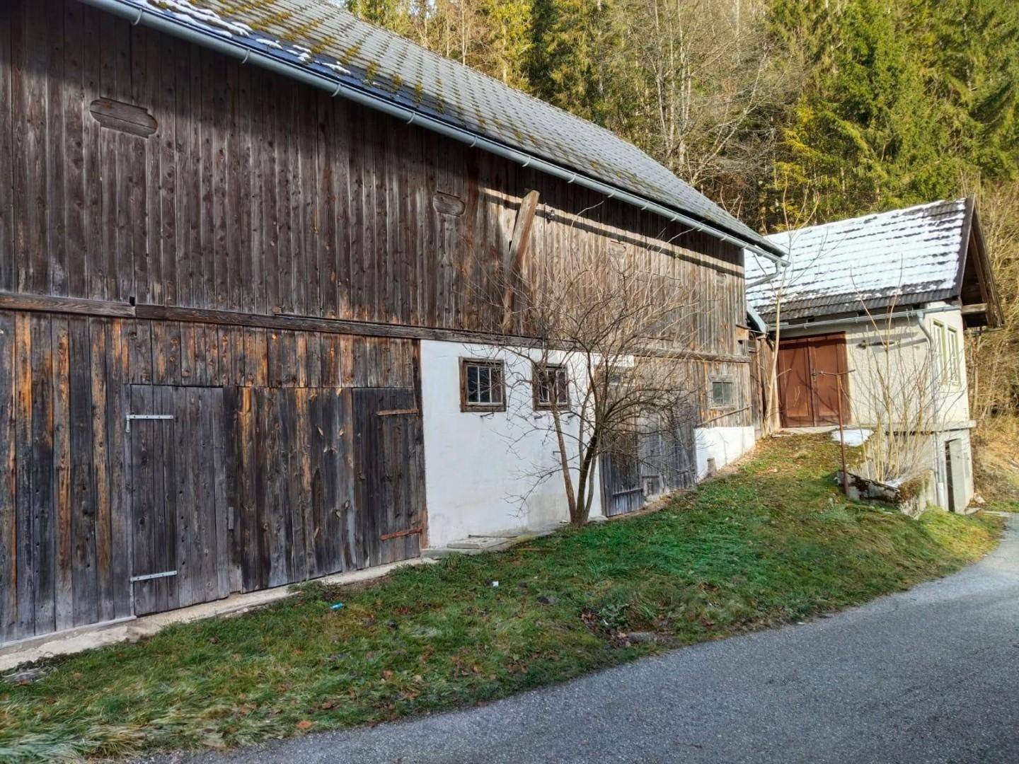 Rustikales Holzgebäude mit großen Scheunentoren und einem kleineren Anbau, umgeben von leichter Vegetation.