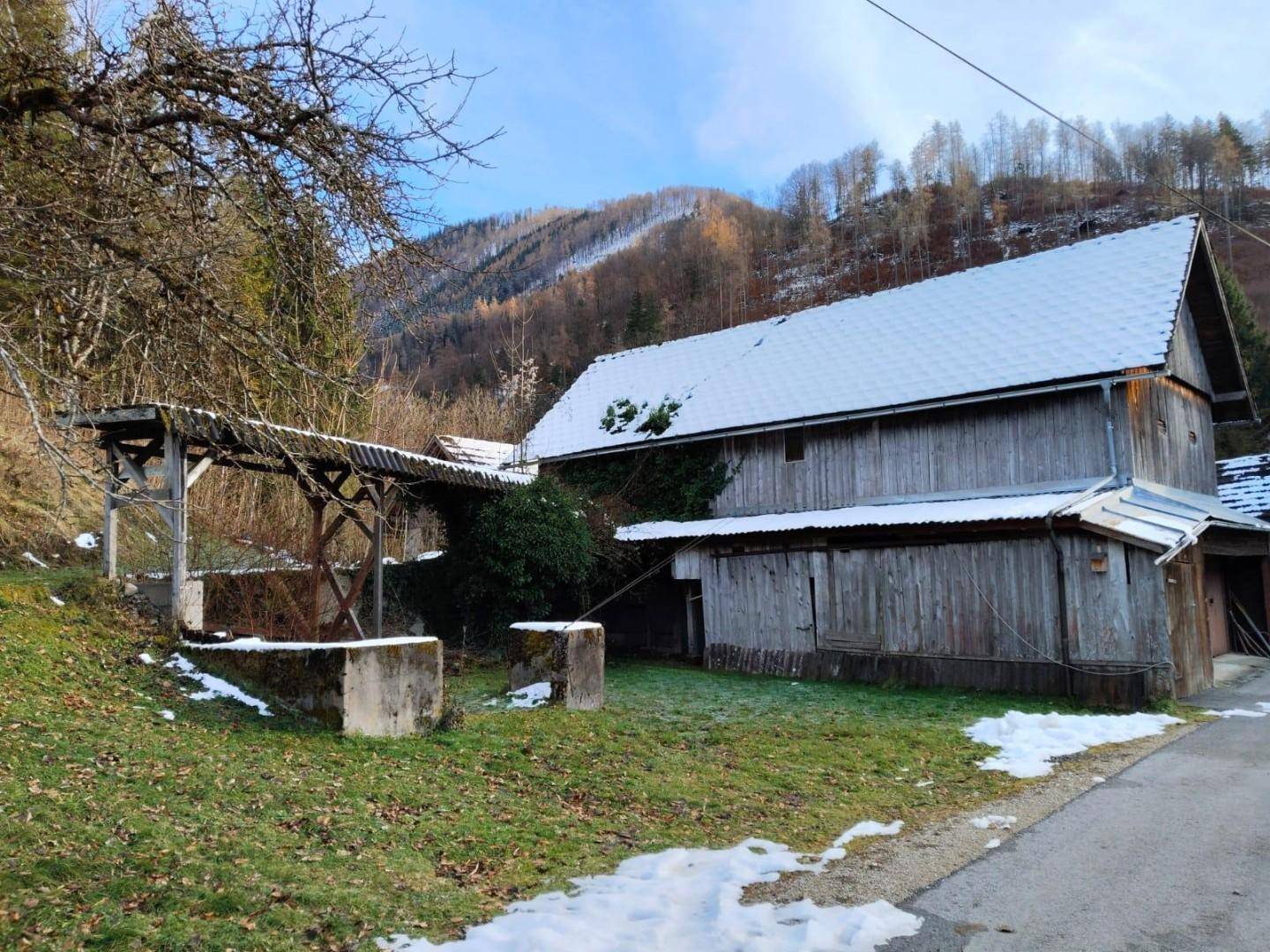 Altes Holzgebäude mit schneebedecktem Dach und einer hölzernen Überdachung, eingebettet in eine ländliche Landschaft.