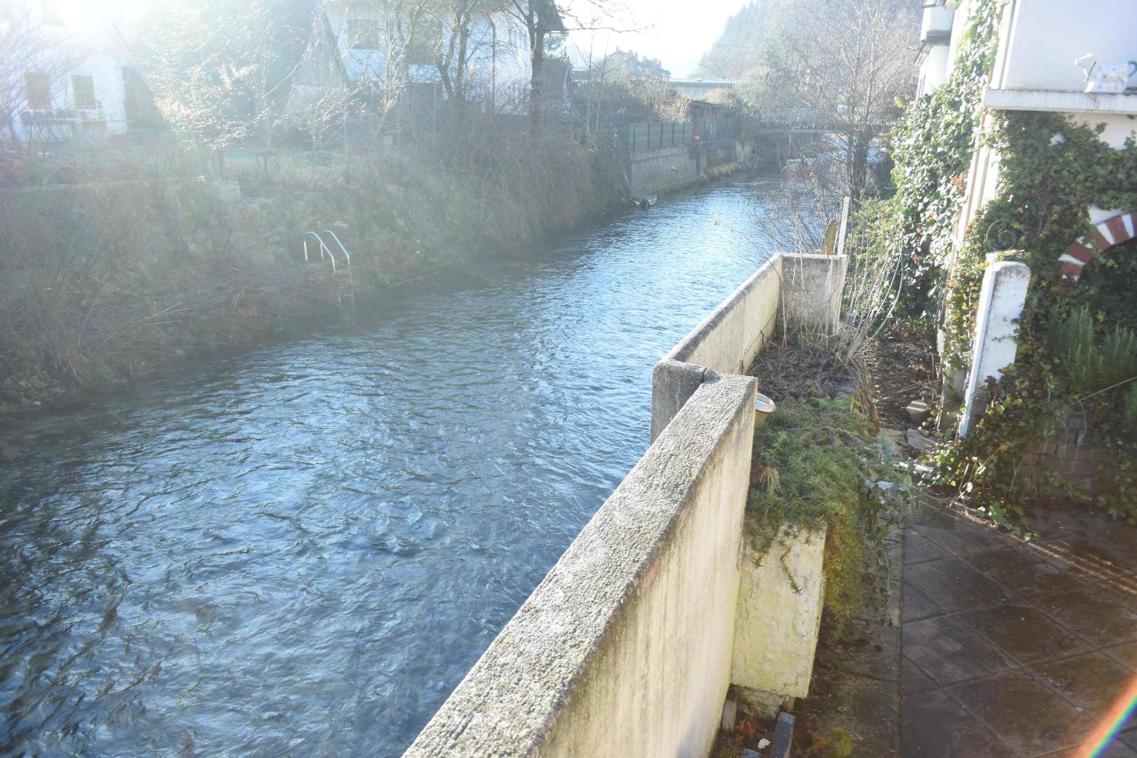 Flusslauf neben dem Grundstück mit Betonmauern und üppiger Vegetation am Ufer.