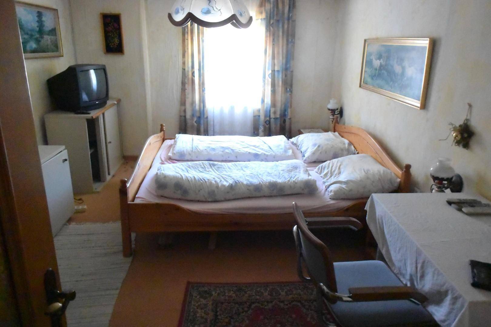 Einfaches Schlafzimmer mit Holzdoppelbett, Teppichboden und Fenster mit Vorhängen.