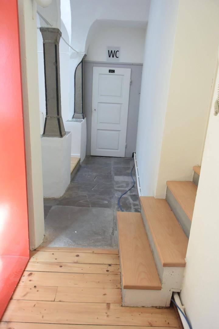 Langer Flur mit Steinfliesenboden, einer Holztreppe und einem WC-Schild über einer weißen Tür, die zu einer Toilette führt.