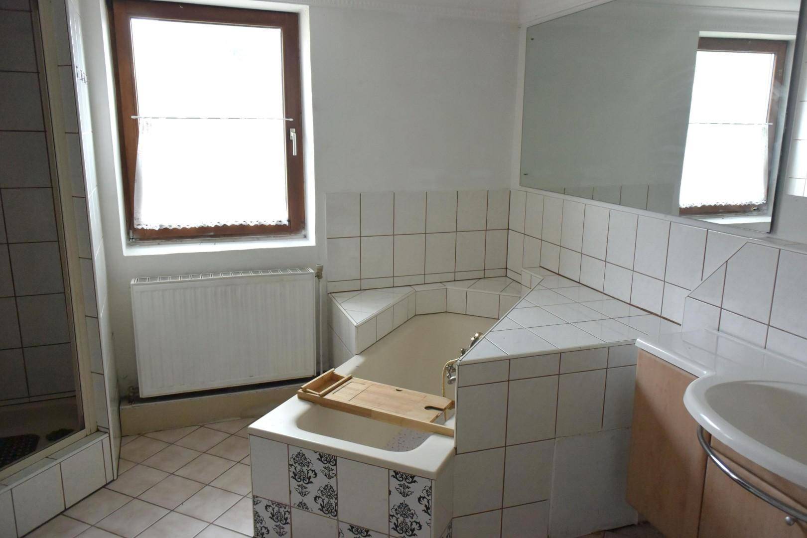 Badezimmer mit Eckbadewanne, weißen Fliesen und einem Fenster für Tageslicht.