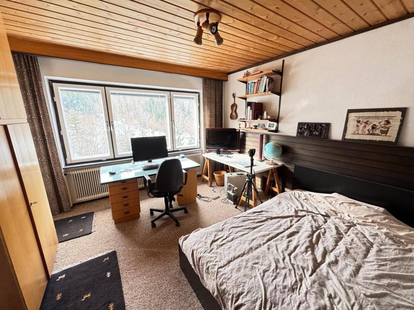 Schlafzimmer mit integriertem Arbeitsbereich, Holzbalkendecke und Blick ins Grüne.