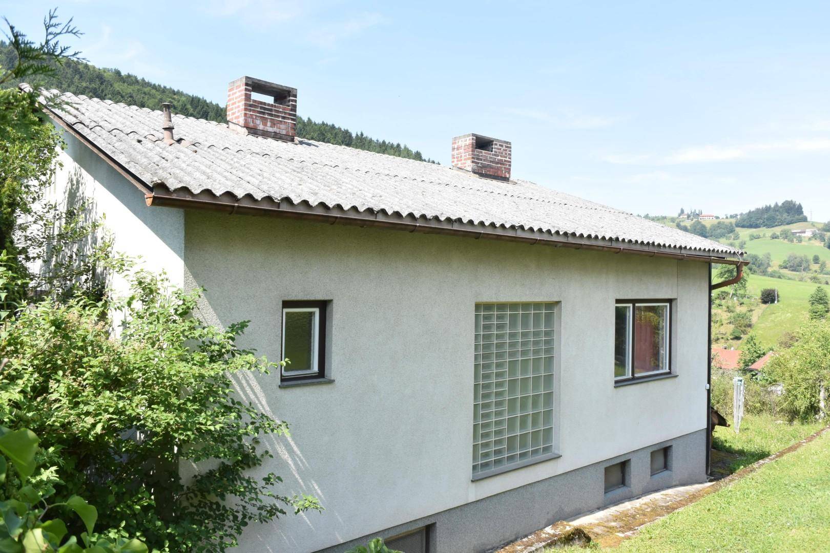 Seitenansicht des Hauses mit Satteldach und Glasbausteinfenster, umgeben von grüner Landschaft.