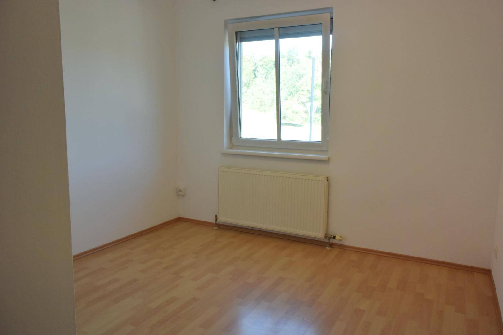 Helles, leeres Schlafzimmer mit Laminatboden, Heizkörper und Fenster.