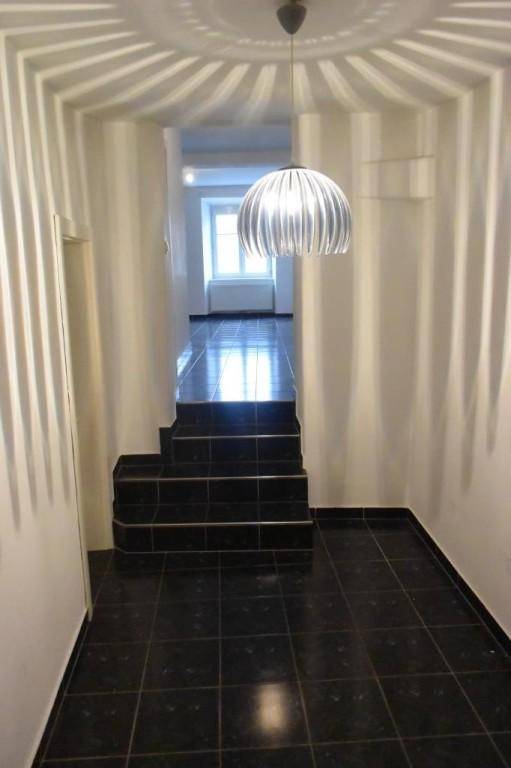 Langer Flur mit dunklen Fliesen und Treppe, beleuchtet von einer modernen Deckenleuchte.