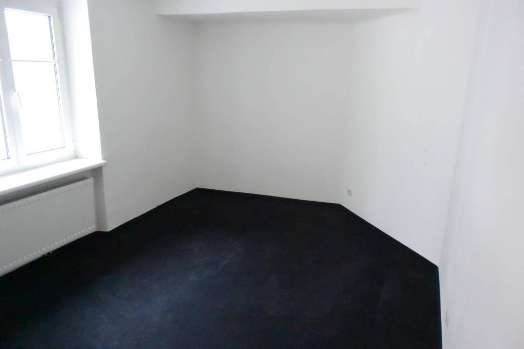 Leerer Raum mit dunklem Boden und weiß gestrichenen Wänden, gut beleuchtet durch ein Fenster.
