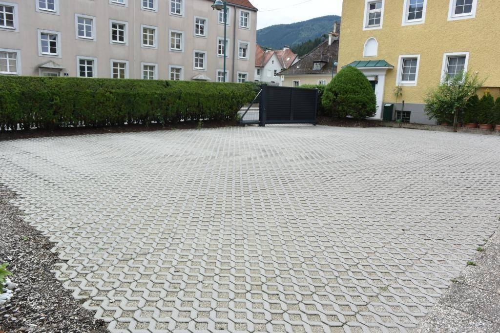 Großer Außenparkplatz mit Rasengittersteinen, umgeben von einer grünen Hecke und Gebäuden.