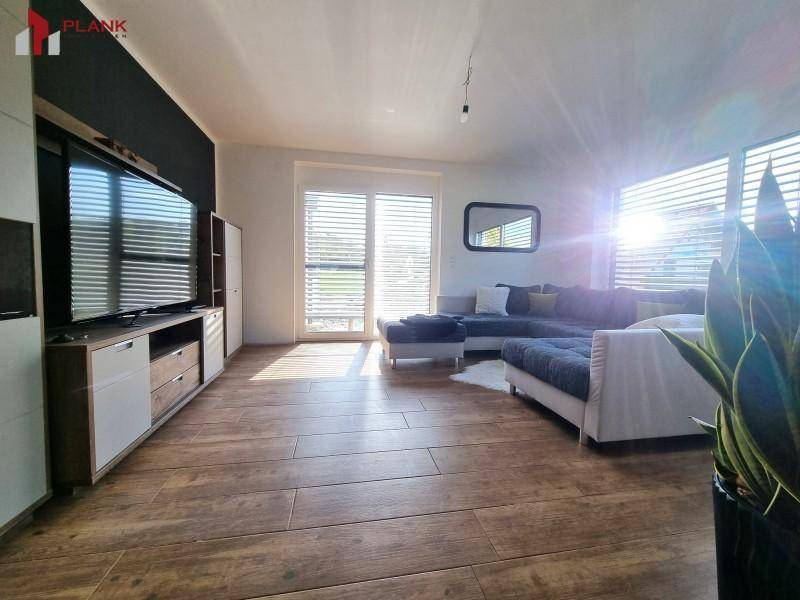 Lichtdurchflutetes Wohnzimmer mit großem Fernseher, Holzboden und Blick ins Grüne.