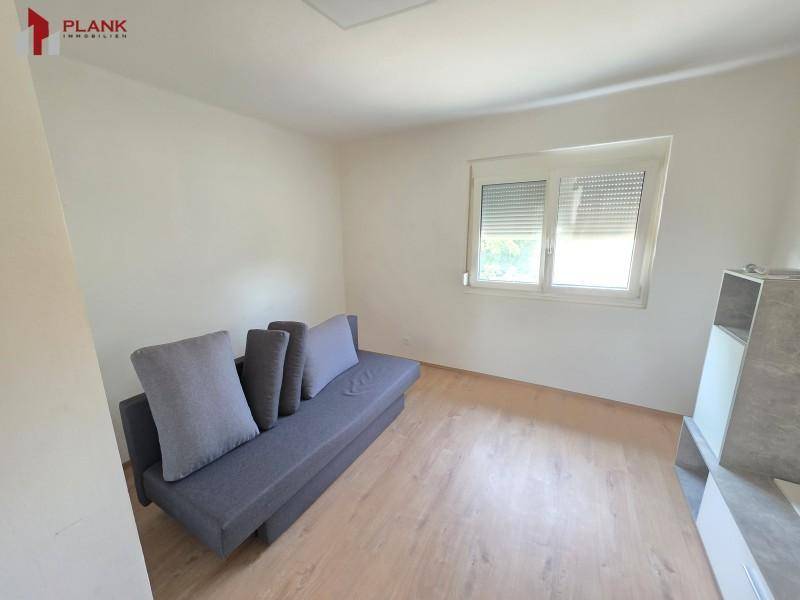 Helles Zimmer mit grauem Sofa, Laminatboden und Fenster mit Blick ins Grüne.