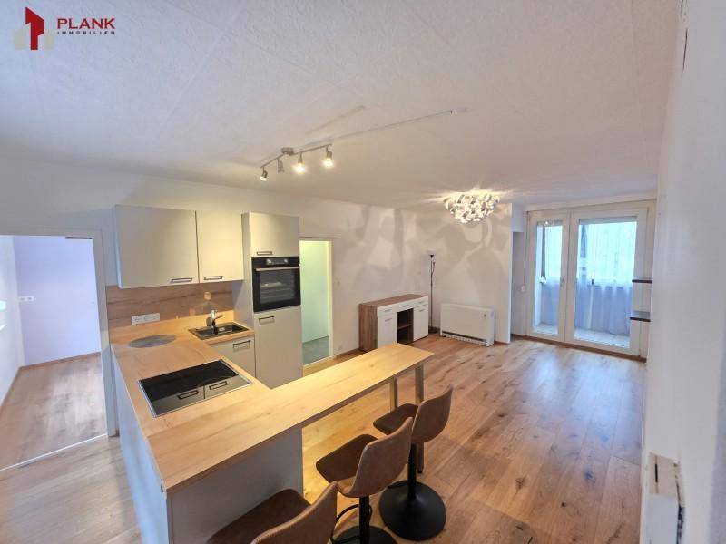 Heller offener Wohnbereich mit moderner Einbauküche, Holzboden und Zugang zum Balkon.