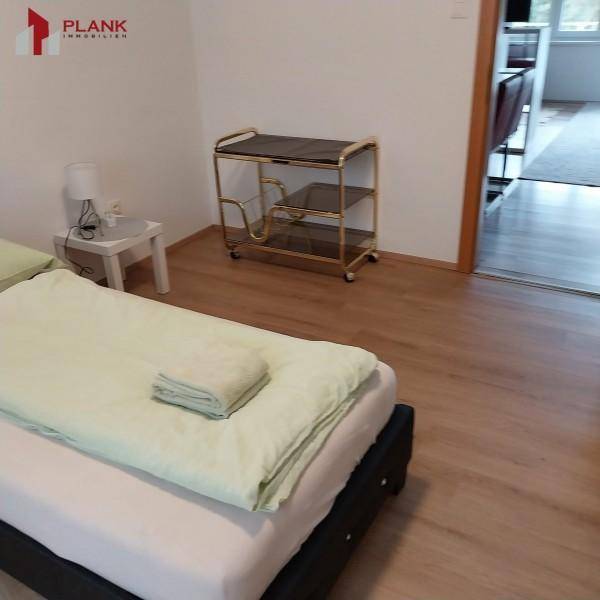 Schlafzimmer mit Einzelbett, Nachttisch und Holzboden, offen zum angrenzenden Wohnbereich.