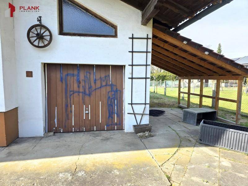 Garagenzufahrt mit Holztor und überdachtem Bereich, umgeben von einem ländlichen Grundstück.