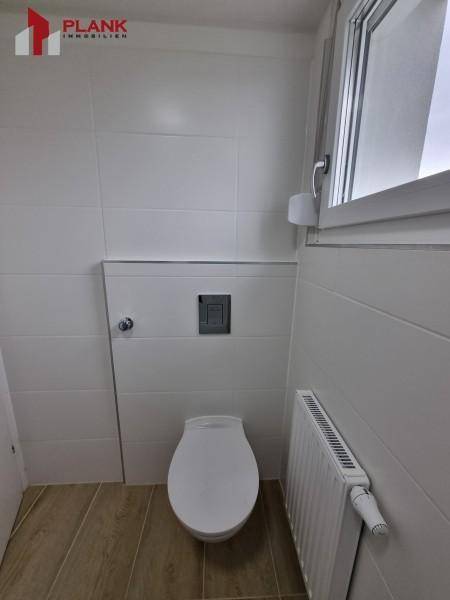 Sauberer Toilettenbereich mit wandhängendem WC, Heizkörper und Fenster für Tageslicht.