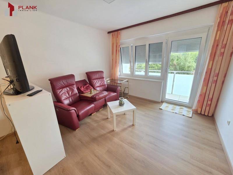 Wohnzimmer mit rotem Ledersofa, Fernseher und Zugang zu einem sonnigen Balkon.