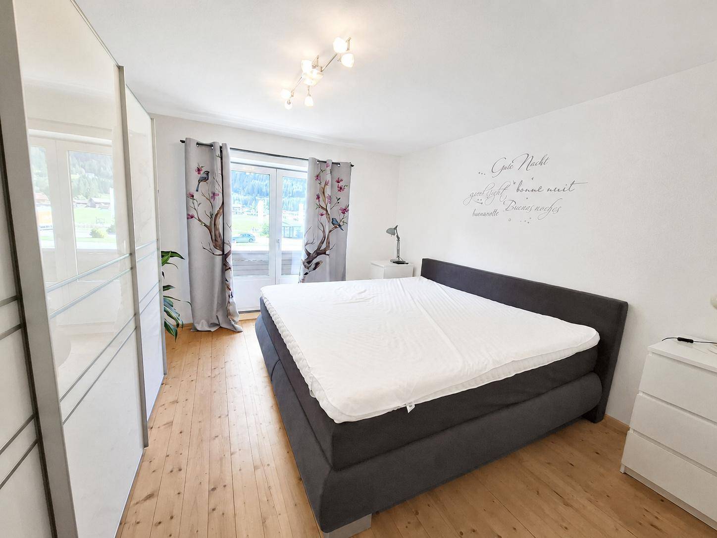 Schlafzimmer mit dunklem Bett, weißem Spiegelschrank, Holzfußboden und Zugang zu einem Außenbereich.