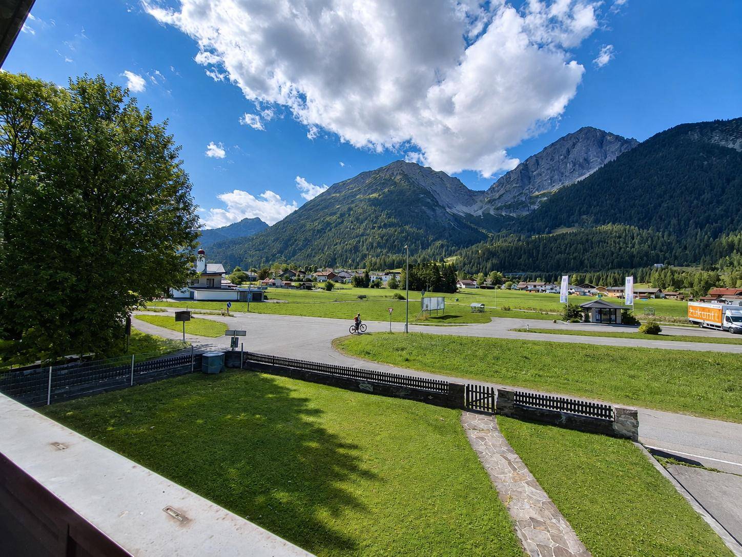 Weitläufiger Blick über grüne Wiesen, eine Straße und majestätische Berge unter blauem Himmel.