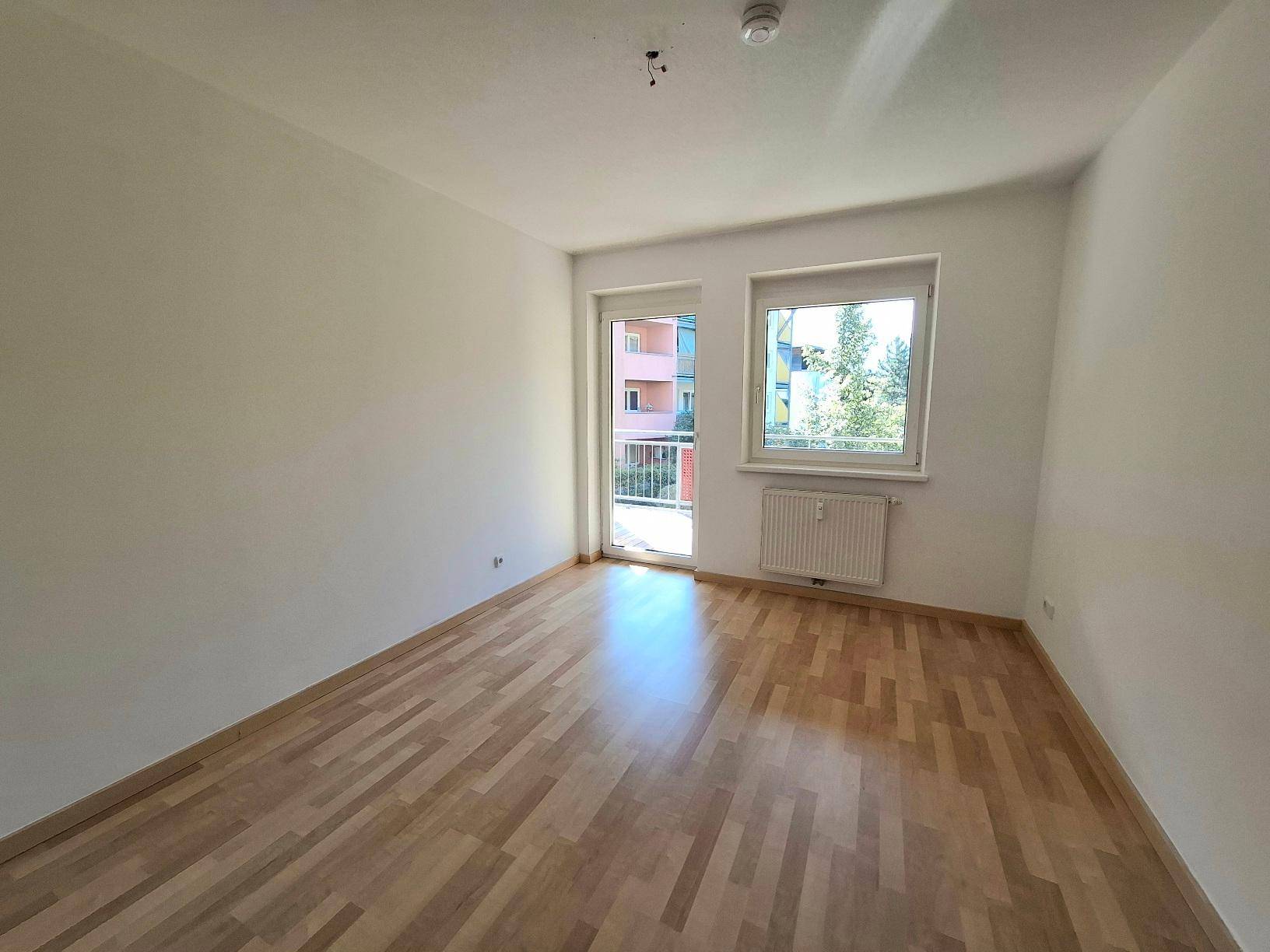 Heller, leerer Raum mit Holzparkettboden, großem Fenster und Zugang zum Balkon.