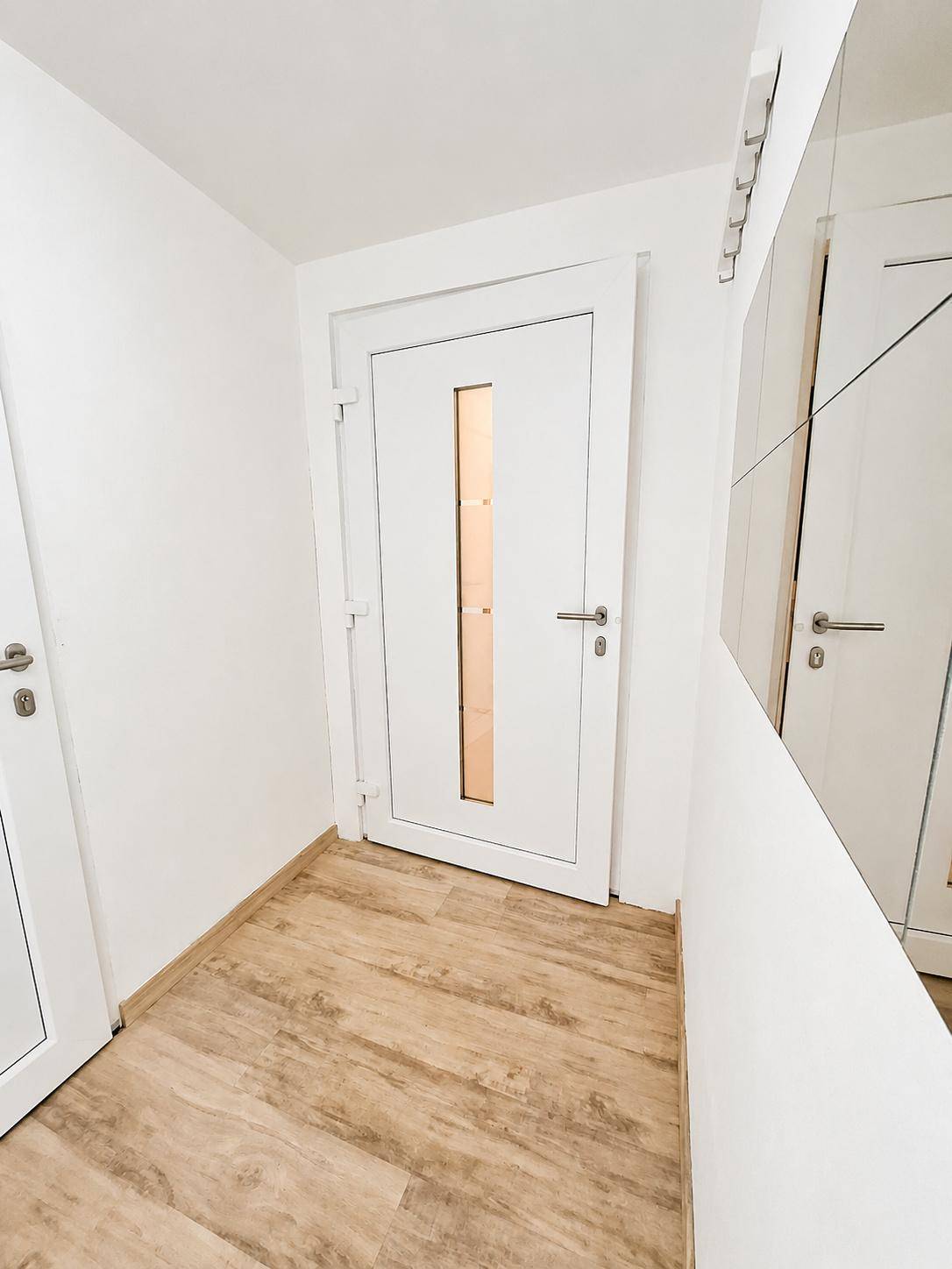 Heller Eingangsbereich mit moderner weißer Tür, Holzfußboden und einer Spiegelfläche an der Wand.